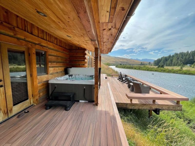 Deck of a four-bedroom Vrbo cabin rental featuring a hot tub and outdoor seating, overlooking the Madison River with mountain views in the distance, near Yellowstone National Park.