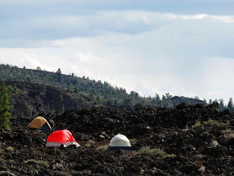 Three tents pitched on the rugged terrain of the Lava Flow Campground in Craters of the Moon National Monument, with lava rocks and a hillside with trees in the background.