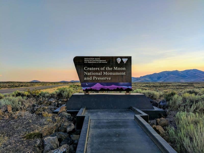 Entrance sign of Craters of the Moon National Monument and Preserve at dusk with the landscape of sagebrush and distant mountains under a gradient evening sky.