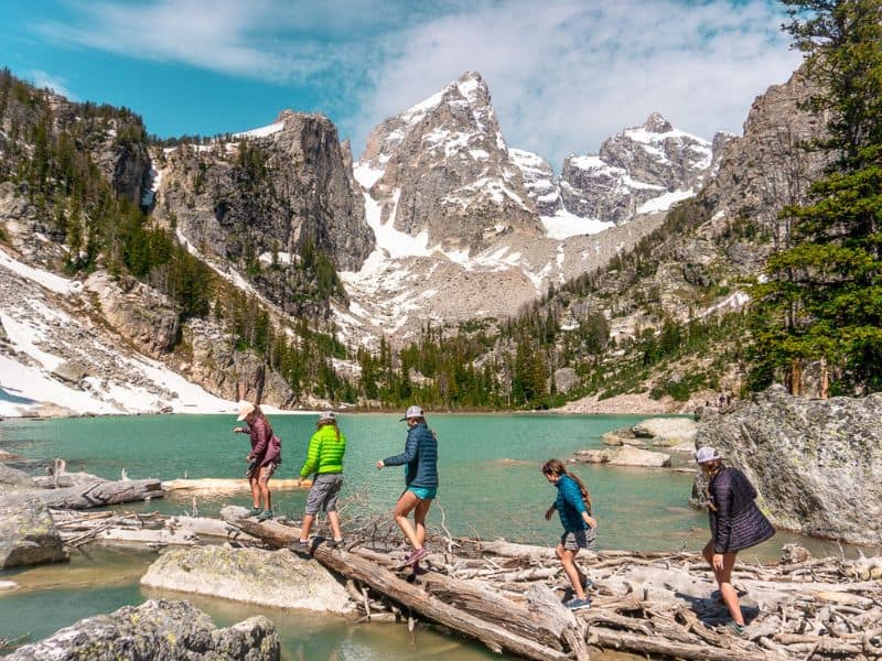 Five hikers cross a log bridge over a turquoise lake surrounded by rugged, snow-capped mountains in Grand Teton National Park. A must see destination on a road trip from Los Angeles to Yellowstone.