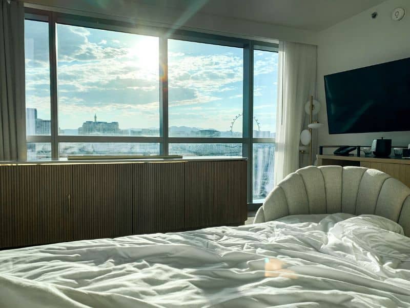 Interior of a hotel room at The Virgin Hotels Resort in Las Vegas with an unmade bed, a curved sofa, and a flat-screen TV, offering a clear view of the Las Vegas Strip through a large window.