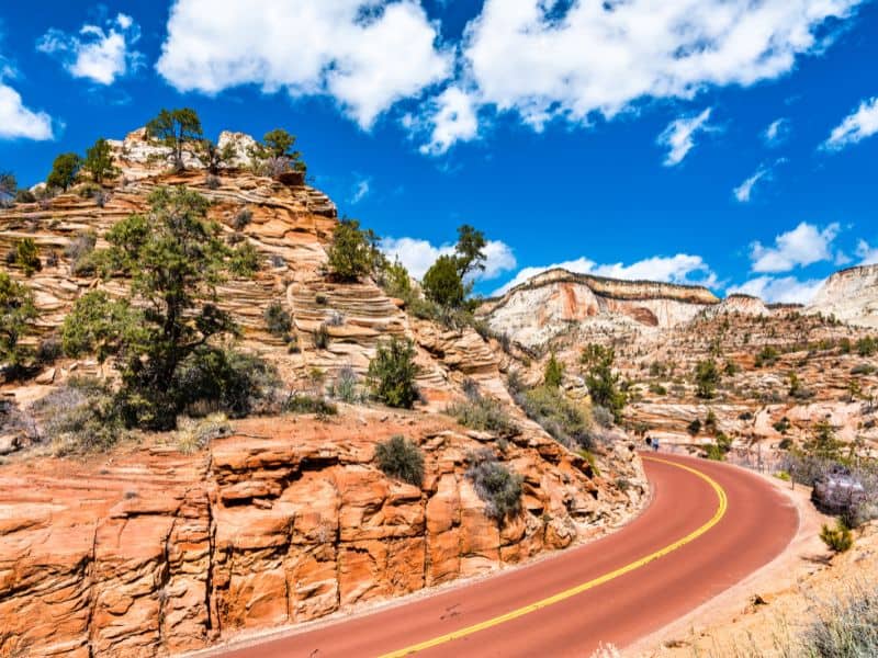 Curved road leading through the red rock formations near Zion National Park under a blue sky with clouds, part of the Los Angeles to Yellowstone road trip itinerary.