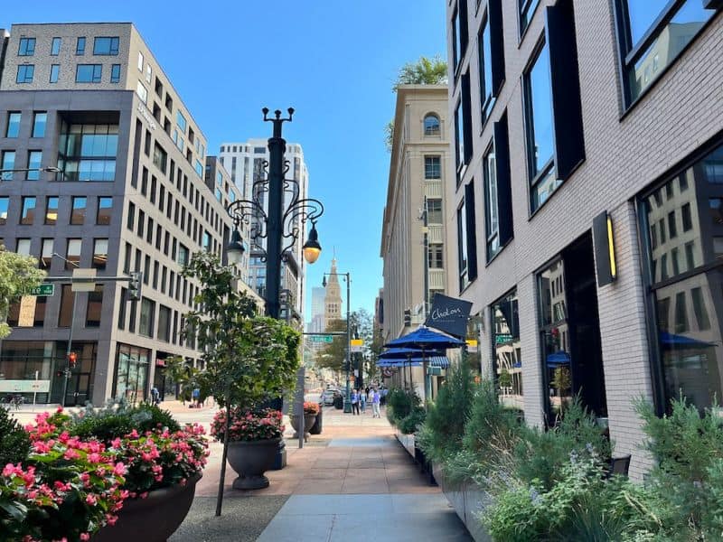 View of 16th Street Mall in Denver, showcasing an urban scene with mixed modern and traditional architecture. The pedestrian street is lined with flowering plants, street lamps, and blue umbrellas. Visible in the distance is the distinctive the Daniels & Fisher Clock Tower.