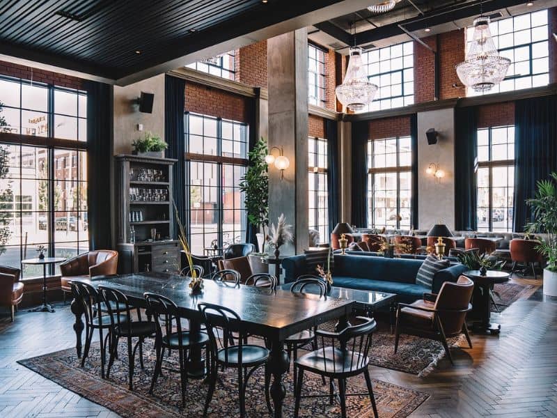 Interior of The Ramble Hotel in Denver featuring a blend of modern and vintage decor. The space includes high ceilings with exposed brick walls, large windows, a long dining table, and multiple seating areas with elegant lighting fixtures.