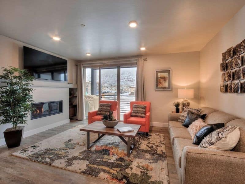 Cozy living room area in Moab Vrbo featuring a modern fireplace, flat-screen TV, and two bright red chairs, looking out to a balcony with a scenic view.