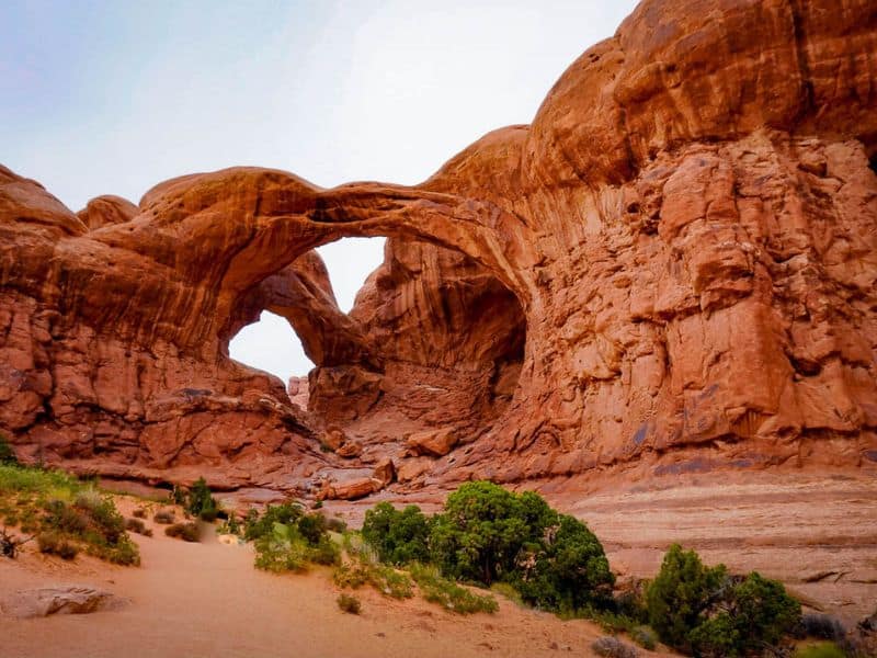 Double Arch at Arches National Park, showcasing two large, connected natural sandstone arches set against a rocky landscape. Small green shrubs and a sandy path are visible in the foreground.