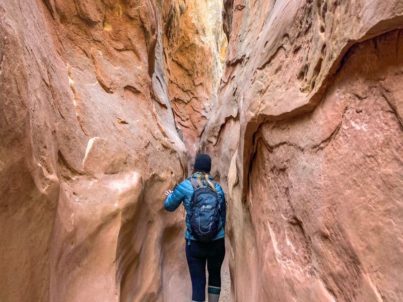 A hiker with a backpack navigates a narrow slot canyon at Little Wild Horse Canyon, with steep, smooth sandstone walls on either side. The canyon walls are reddish-brown, and the path is tight and winding.