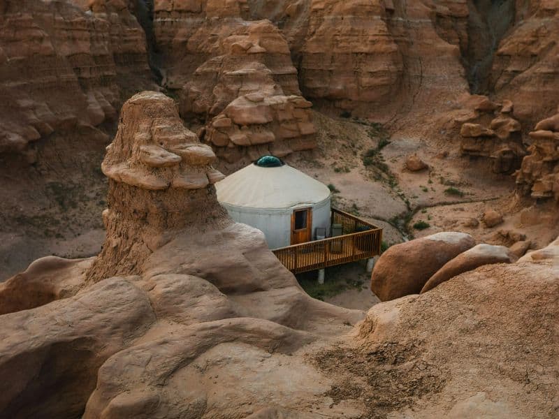 Yurt nestled among rock formations at Goblin Valley State Park, featuring a wooden deck and surrounded by unique, eroded sandstone hoodoos. The landscape is rugged and rocky.