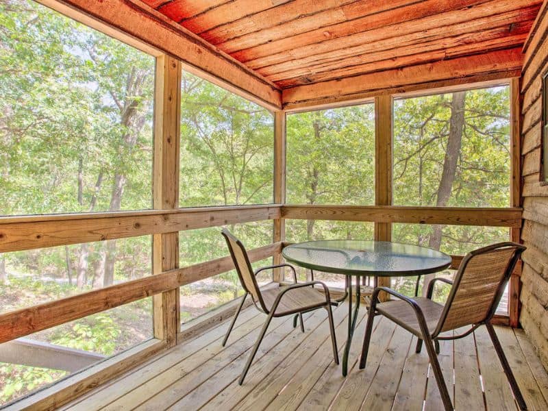 Screened-in porch at Bright Davis Escape studi cabin near Turner Falls, with a round glass table with two chairs and a view of the surrounding trees.