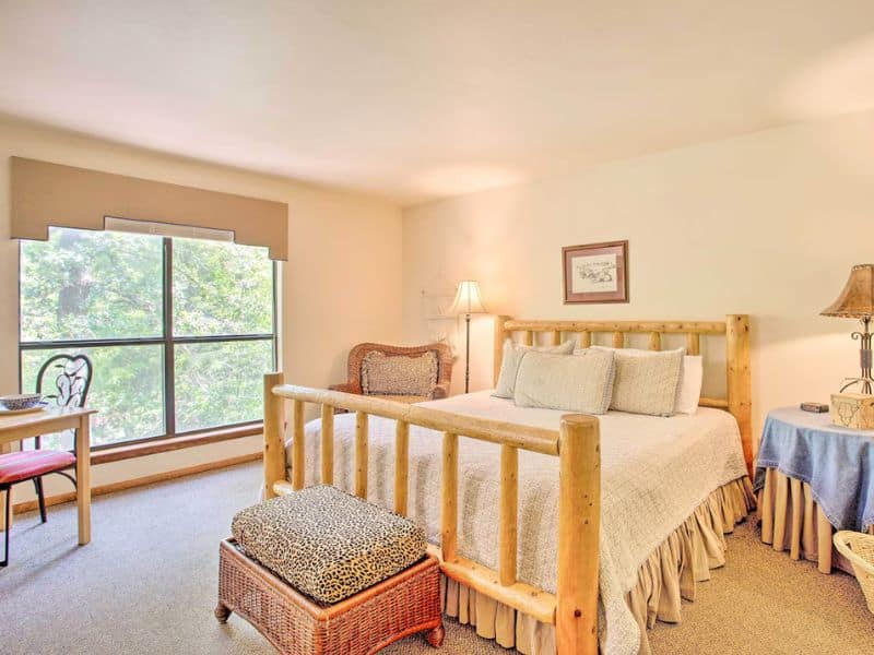 Studio bedroom inside Bright Davis Escape cabin rental near Turner Falls, showcasing a wooden bed frame, a large window, a wicker chair, a bedside table with a lamp, and a small writing desk.