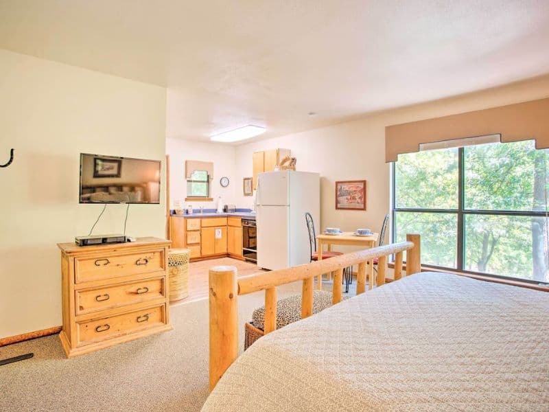 Combined bedroom and kitchen area of Bright Davis Escape Vrbo cabin studio, including a bed, a TV on a dresser, a table with chairs by the window, and a small kitchen with a refrigerator and stove.