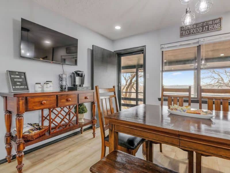 Dining area in Hickory Hill Hideaway cabin near Turner Falls with a wooden table and chairs, a sideboard with a coffee maker and decor, a wall-mounted TV, and large windows with a view outside.