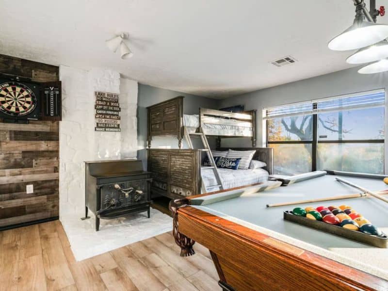 Recreation room in a Turner Falls cabin rental featuring a pool table, dartboard, bunk beds, a wood burning stove, and large windows offering a scenic view.