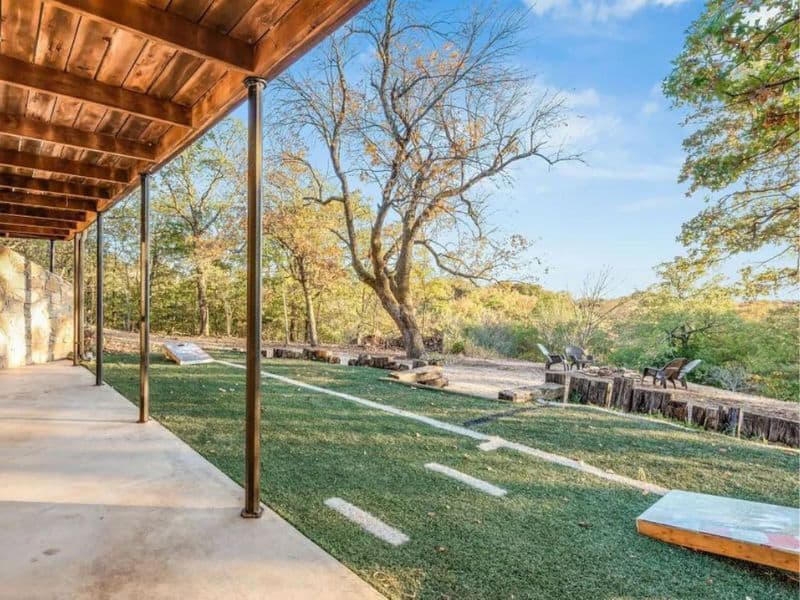Outdoor area at a Vrbo rental near Turner Falls with a covered patio, a grassy yard with cornhole boards, and a fire pit area surrounded by trees.