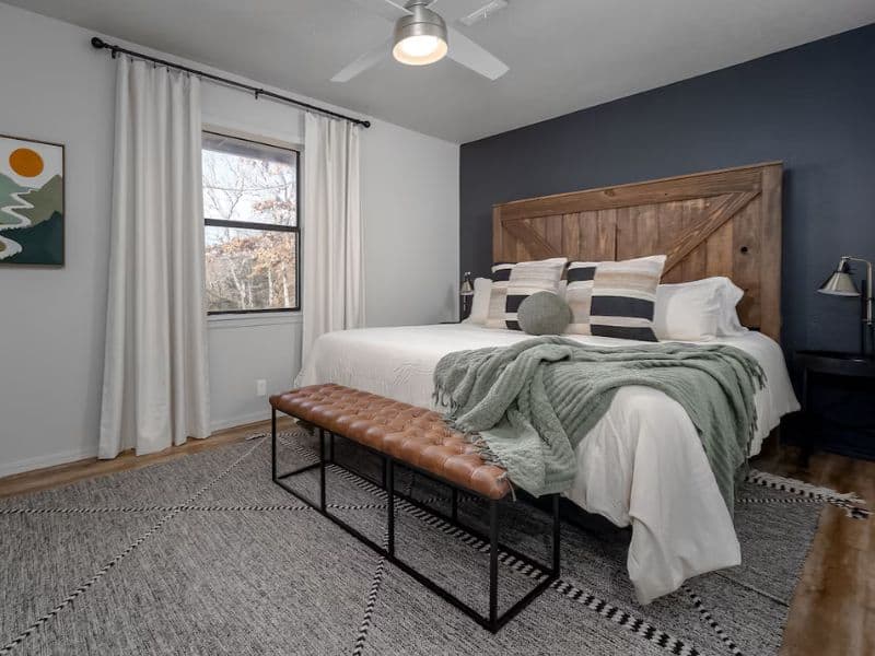 Bedroom inside a Turner Falls cabin rental with a large bed with pillows and a green throw, a wooden headboard, a leather bench at the foot of the bed, a ceiling fan, and a window with white curtains.
