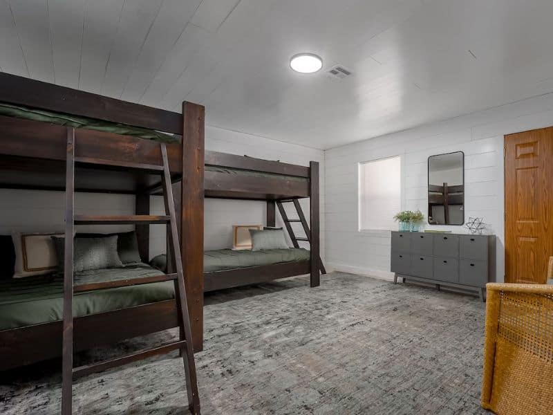 Bunk bed room in a cabin rental near Turner Falls, featuring two sets of dark wooden bunk beds with green bedding, a gray dresser, a mirror, and a small window, all in a white-paneled room.