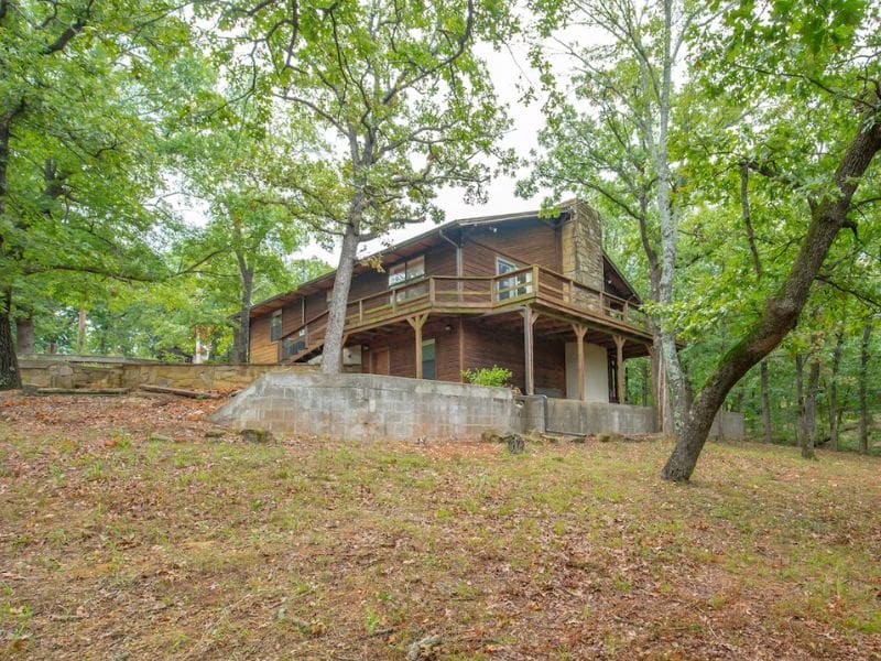 Exterior of a Turner Falls cabin rental surrounded by trees, showing a wooden deck and a stone foundation, with the cabin situated on a slight hill.