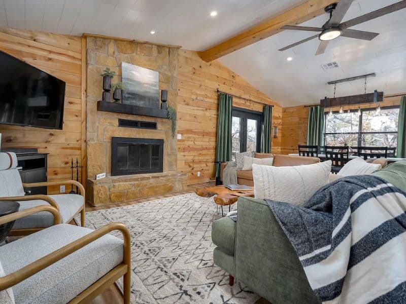 Interior of a cabin featuring a living room with a stone fireplace, a mounted TV, wooden walls, and modern furniture, including a green couch, chairs, and a patterned rug. Cabin rental near Turner Falls, OK.