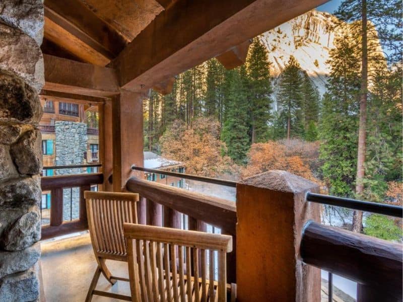 Balcony view from the Ahwahnee Hotel in Yosemite Valley, with wooden chairs and a scenic backdrop of tall evergreen trees and towering granite cliffs.