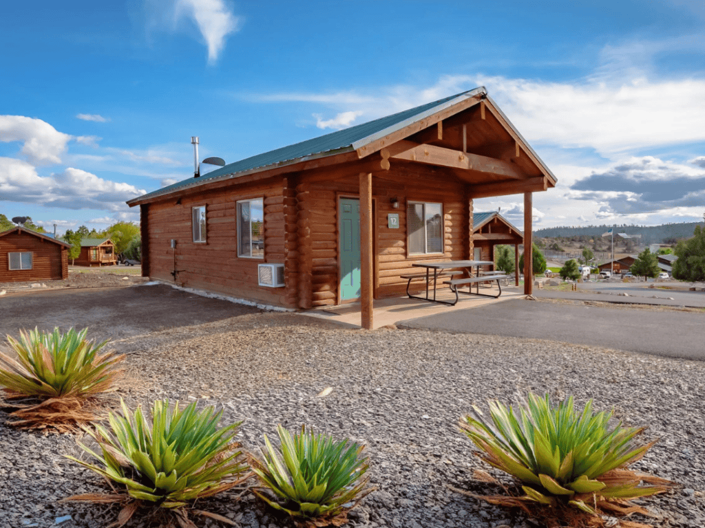 Exterior of a rustic log cabin at Zion Ponderosa Ranch Resort with a green roof and a picnic table outside. The cabin is surrounded by desert landscape and a clear blue sky.
