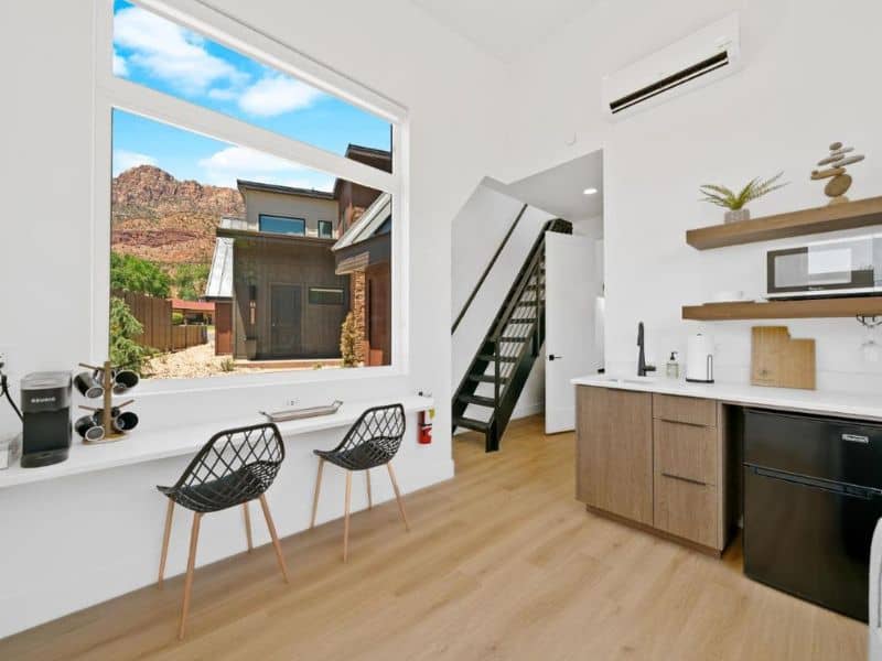 Interior view of a modern bungalow at The Bungalows at Zion with a bright, spacious living area. The room features a large window with a view of the red rock formations outside, two stylish chairs, and a small kitchenette with wooden cabinets.
