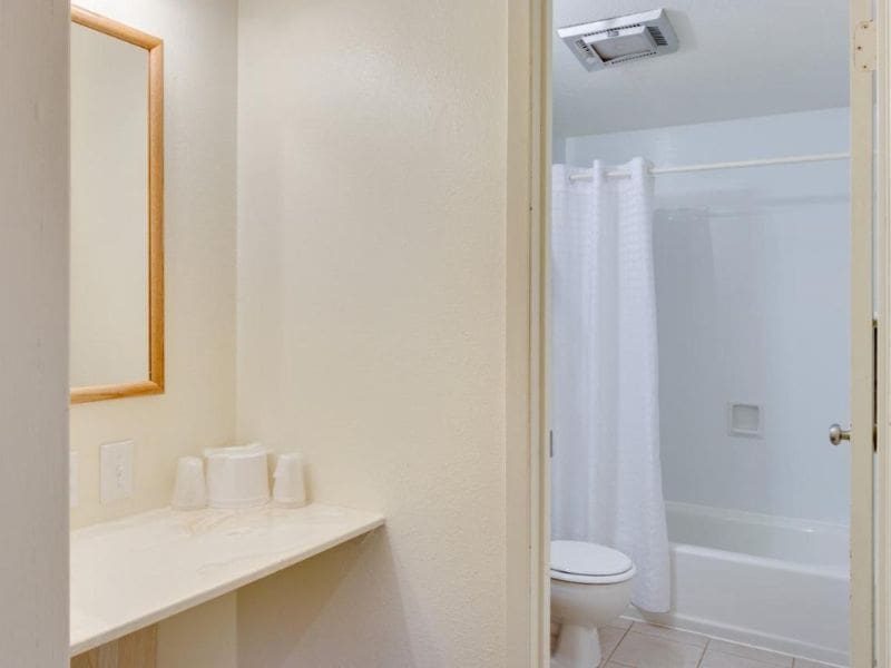 Simple and clean bathroom setup with a vanity area, a mirror, and a sink, adjacent to a white-tiled bathtub and shower with a white shower curtain.