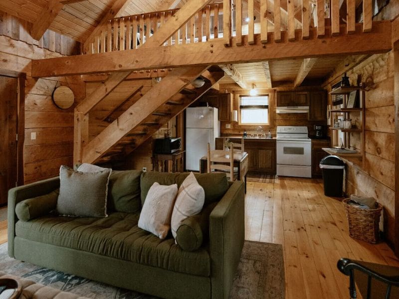 A cozy living area featuring a lofted wooden staircase, a plush green couch, and a small kitchen area with rustic wood interiors. Pet-friendly cabin for 2 in Hocking Hills, Ohio.