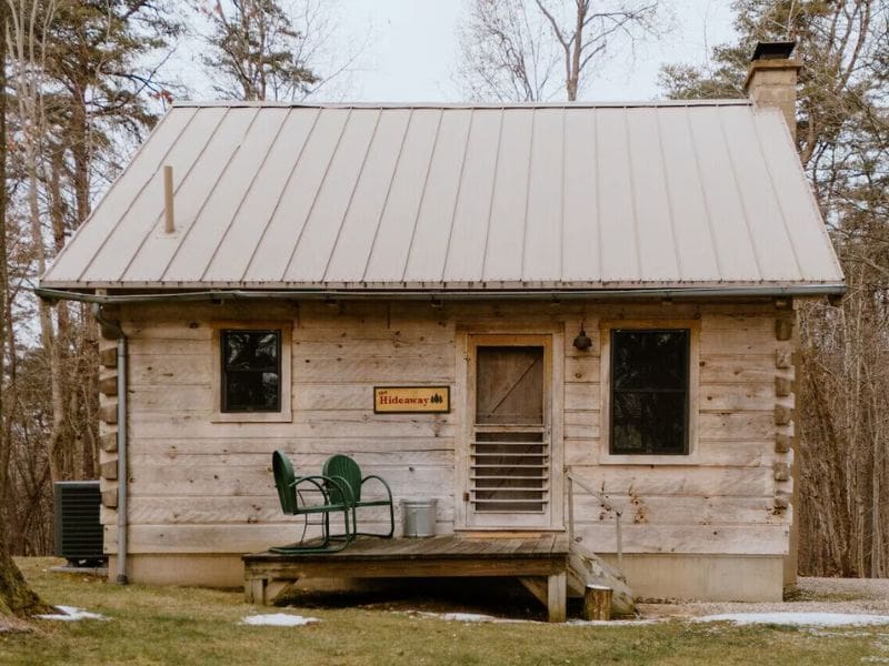 A cute rustic cabin with a metal roof and a small front porch set among the trees with a sign reading "Hideaway." A dog-friendly cabin for two in Hocking Hills, OH.